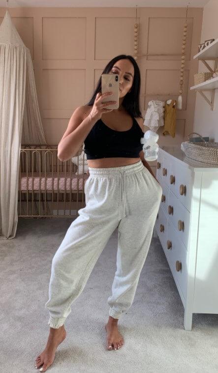 A mother taking a mirror selfie in a nursery, wearing a nursing top and sweatpants, holding a Momcozy hands-free breast pump.