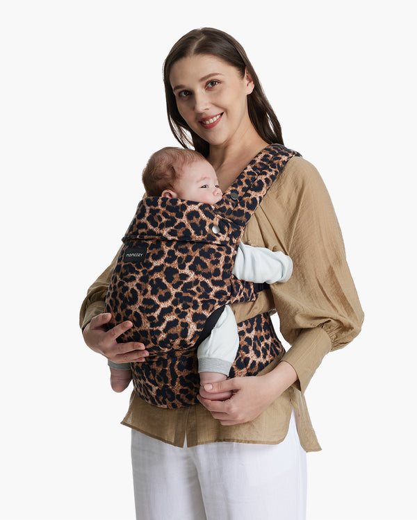 Smiling woman wearing a beige blouse and white pants carrying a baby in a Momcozy leopard print baby carrier