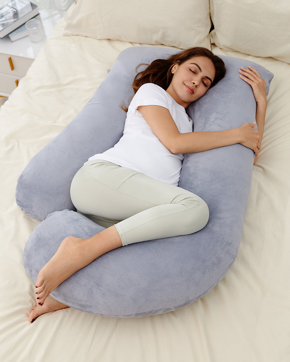 Pregnant woman resting on a U-shaped maternity pillow for comfortable sleep and body support, light gray plush material.